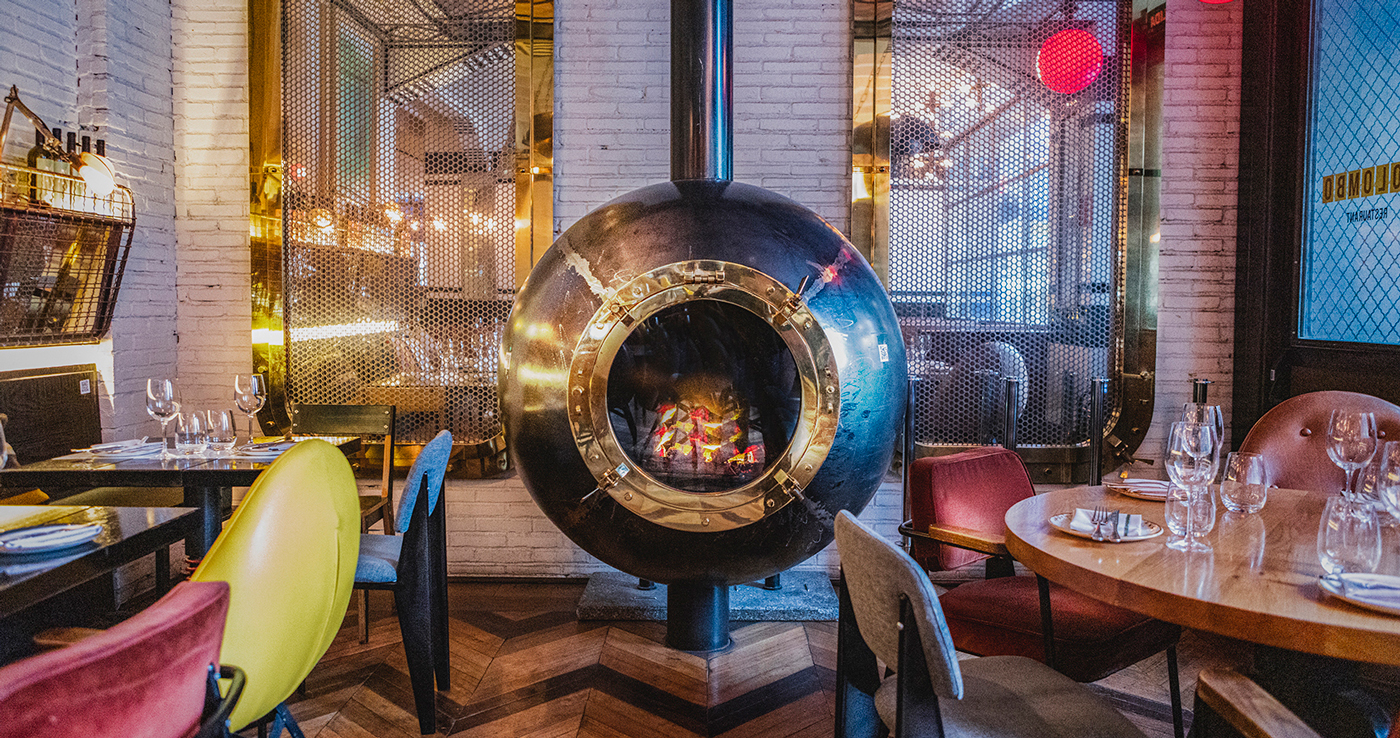 Plano del restaurante Colombo en el que se ve decoración de estilo industrial con ladrillos, metal…En los laterales de la imagen hay mesas con sillas cómodas y copas de cristal. En el centro de la imagen hay un gran horno de metal redondo.